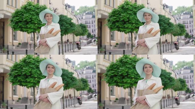 戴着优雅帽子的时尚女士站在城市街道上，戴着法式长棍面包。法国-在阳光明媚的夏日在欧洲旅行的成年白人妇
