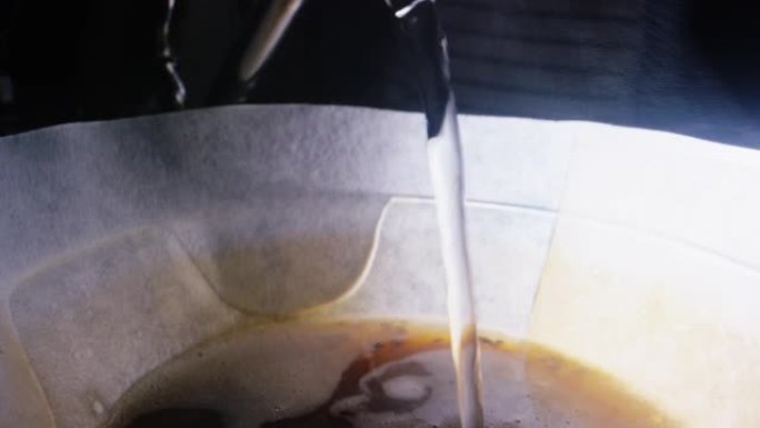 从金属茶壶中喷口的宏观拍摄将热水倒入带有咖啡渣的纸过滤器中，并在咖啡店的玻璃倒咖啡壶中倒入咖啡渣
