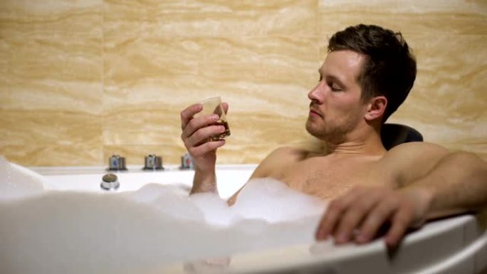 心满意足的男人喝着浓威士忌躺在有泡沫的浴缸里，放松