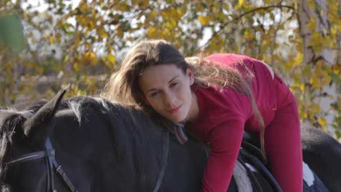 年轻快乐的高加索女性马术者躺在马背上爱抚她的宠物。美丽的黑马站在秋天的森林中，年轻的骑手在他的背上。