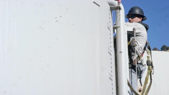 一个阳光明媚的早晨，一名二十多岁的男性油田工人在油气钻井平台现场的泥浆罐侧面的梯子顶部的手持收音机上