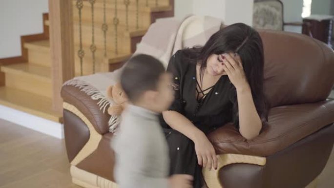 疲惫的中东母亲坐在扶手椅上，她的两个小孩跑来跑去
