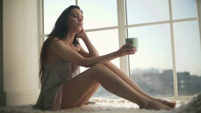 穿着睡衣的漂亮女孩从地板到天花板坐在窗户旁边。穿着睡衣的年轻女子喝着早上的咖啡，看着窗外。