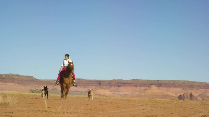 在晴朗明亮的日子里，一个穿着传统纳瓦霍族服装的十几岁的美国原住民女孩骑着她的马穿过纪念碑谷沙漠，带着