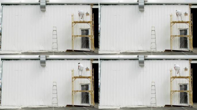 一位三十多岁的高加索专业画家在部分多云的天空下站在脚手架上时，使用喷漆器对金属仓库的外部进行油漆