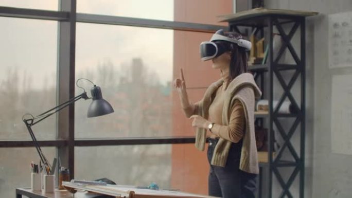 办公室里的现代女性建筑师带着大窗户站在虚拟现实头盔上，不用离开办公室就可以使用手势来管理项目。施工控