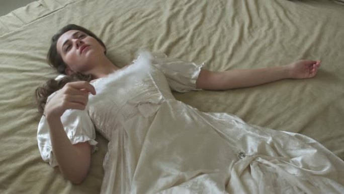 穿着白色复古舞会礼服的年轻美女躺在巨大的金色床上，挥舞着羽毛风扇。