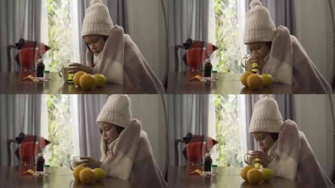 穿着冬帽围巾的小女孩的特写镜头被毯子盖着，坐在桌旁喝茶