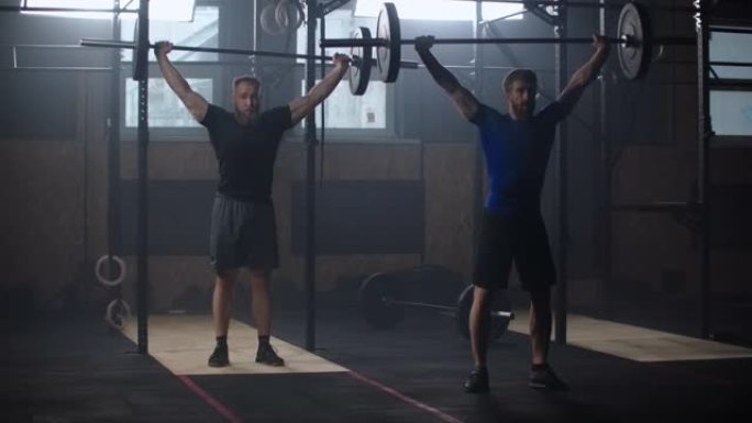 两个强壮的男人在健身房一起做举重训练。中等射击慢动作平底锅男子在健身房用运动器材锻炼: 头顶深蹲、平