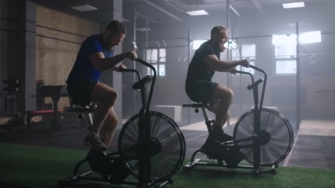 2个年轻的活跃男子在健身房锻炼空气自行车。男性在慢动作中进行空中自行车训练。中等身材的健身男子在复制