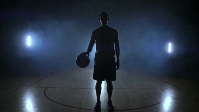 篮球运动员站在黑暗的操场上，将球握在手中，在黑暗中看着相机，慢动作的背光和烟雾笼罩着