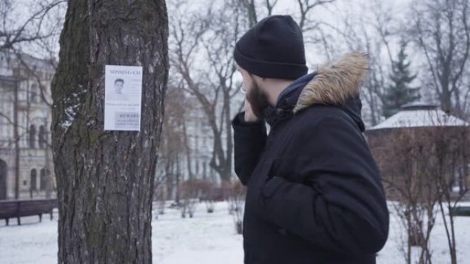 年轻的大胡子高加索人在电话里聊天，看着挂在树上的失踪人员广告。穿着冬装的移情家伙帮助人们寻找失散的人