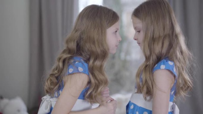 愤怒的高加索小女孩责骂她的双胞胎妹妹的侧视图中间镜头。穿着蓝色圆点连衣裙的漂亮兄弟姐妹在室内争吵。沟