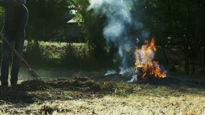一个四十多岁的高加索人用干草叉拿起木棍和树枝，走向户外的一个大火烧堆
