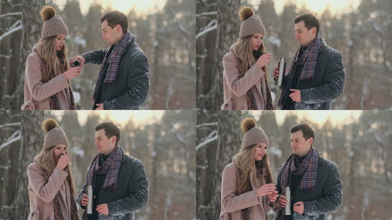 恋爱中的情侣在冬季森林里喝热水瓶里的茶。冬天在公园里穿着外套的时尚男人和女人散步