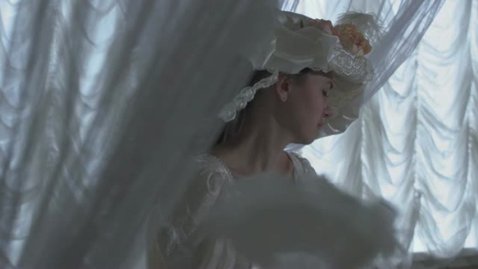 穿着白色复古舞会礼服和白色帽子的漂亮年轻女子站在透明窗帘和挥舞风扇的背景上