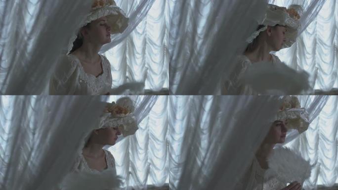 穿着白色复古舞会礼服和白色帽子的漂亮年轻女子站在透明窗帘和挥舞风扇的背景上