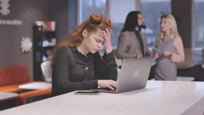 疲惫的红发白人妇女在笔记本电脑键盘上打字并用手握着头的肖像。疲惫的女员工在开放空间办公室过度工作。电
