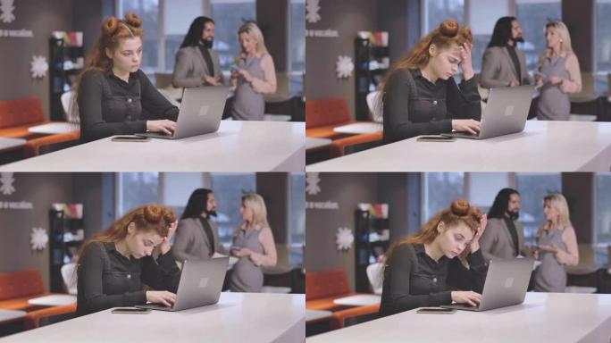 疲惫的红发白人妇女在笔记本电脑键盘上打字并用手握着头的肖像。疲惫的女员工在开放空间办公室过度工作。电