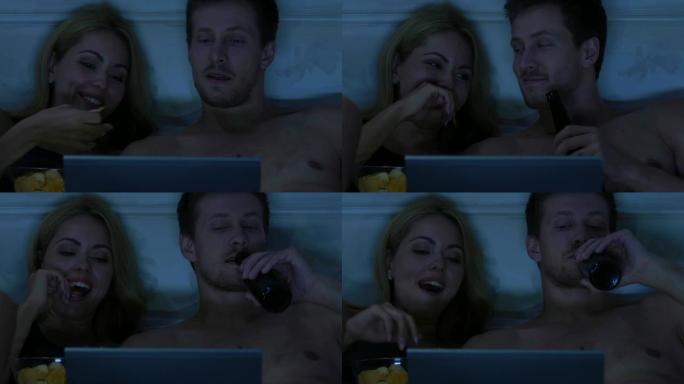 年轻夫妇在床上看笔记本电脑上的连续剧，吃零食和喝啤酒