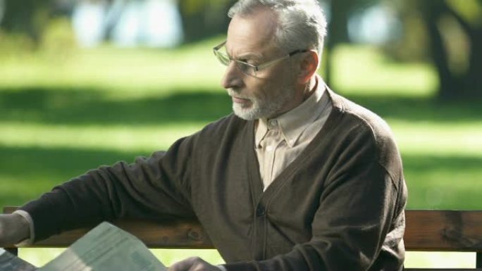 戴眼镜的退休男子读报纸，早上坐在公园长椅上