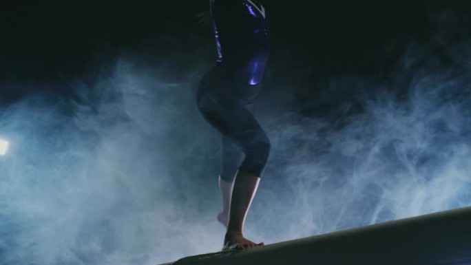 女孩体操运动员的腿的特写镜头在烟雾和慢动作中进行平衡木跳跃