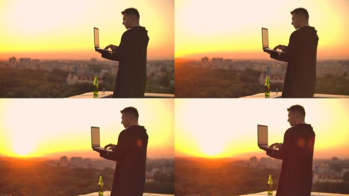 日落时分站在屋顶上拿着笔记本电脑和啤酒。一名身穿帽衫的男子在高处欣赏着美丽的城市风景