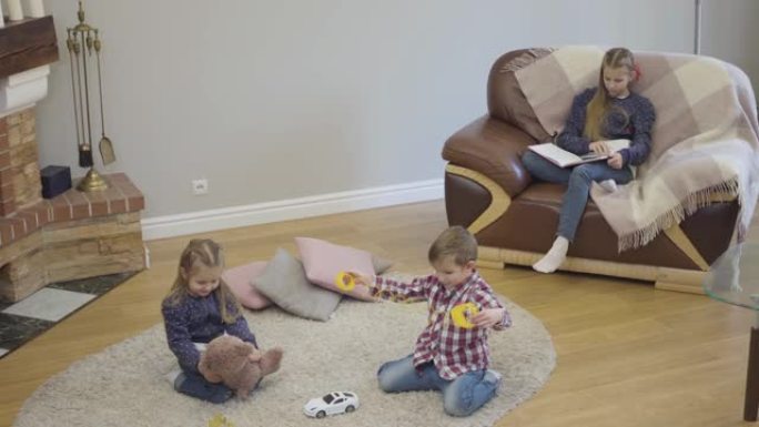 欢快的高加索兄弟姐妹在家里玩耍的俯视图，作为他们的姐姐看书。平静的孩子在家里一起度过空闲时间。休闲，