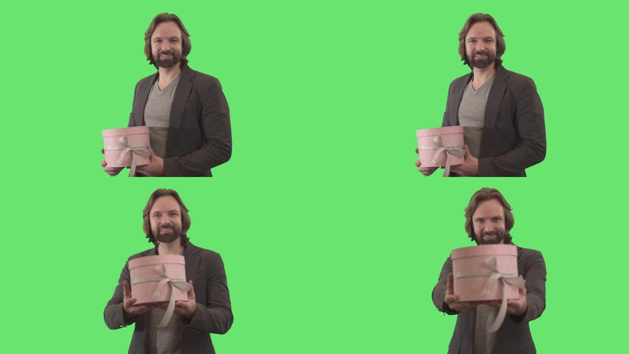 微笑的黑发白人男子将礼品盒伸向相机。留着胡子的成年男子的肖像，在绿色背景下摆着粉红色的礼物。节日，庆