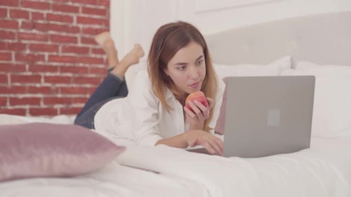 年轻的白人妇女躺在床上在笔记本电脑上打字并吃红苹果