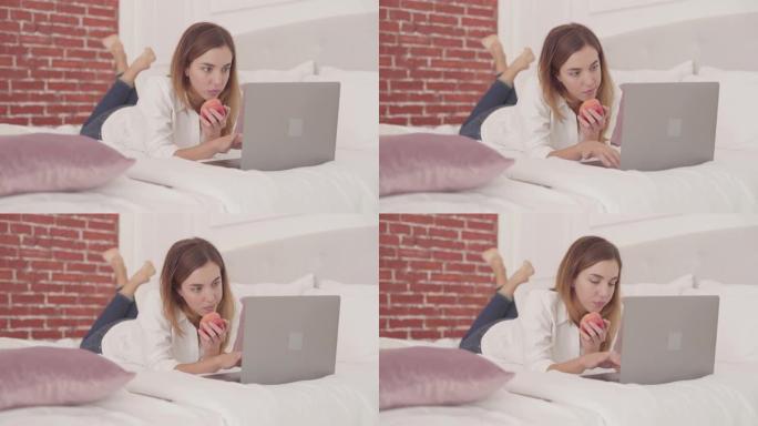 年轻的白人妇女躺在床上在笔记本电脑上打字并吃红苹果