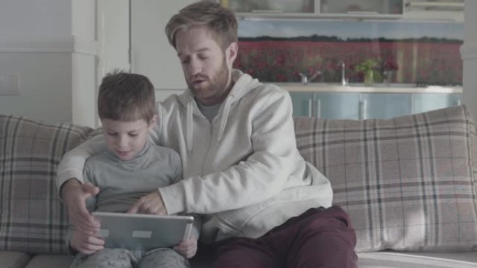 父子俩使用数字平板电脑坐在大客厅的沙发上。爸爸教他的孩子。幸福的家庭在一起。父子关系。