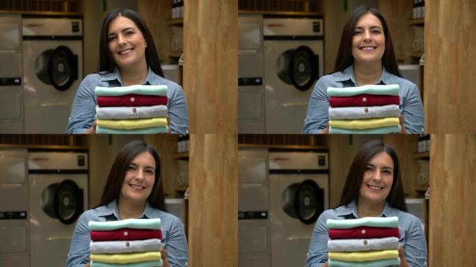 开朗的成熟女性顾客在自助洗衣店拿着一叠折叠的衬衫，对着镜头微笑
