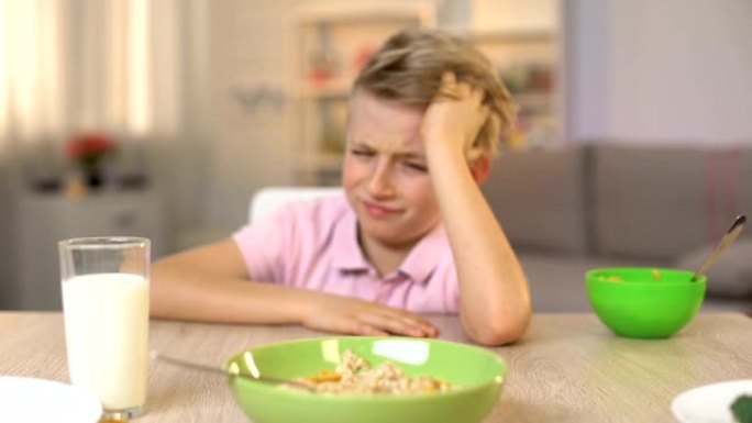 不快乐的男孩看着燕麦片，厌恶，令人垂涎的食物，健康的早餐