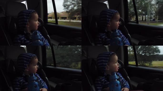 一个穿着连帽衫的三岁白人男孩坐在他的汽车座位上，看着行驶中的车辆的窗外