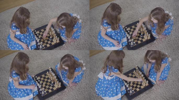 两个黑发高加索双胞胎姐妹在室内下棋的俯视图。穿着优雅蓝色连衣裙的小女孩在家一起休息。智力，爱好，生活
