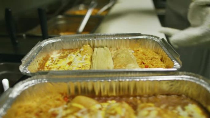 一个人在墨西哥餐厅的商业厨房里，将未包装的玉米粉蒸肉放入带有奶酪和米饭的金属烤盘中