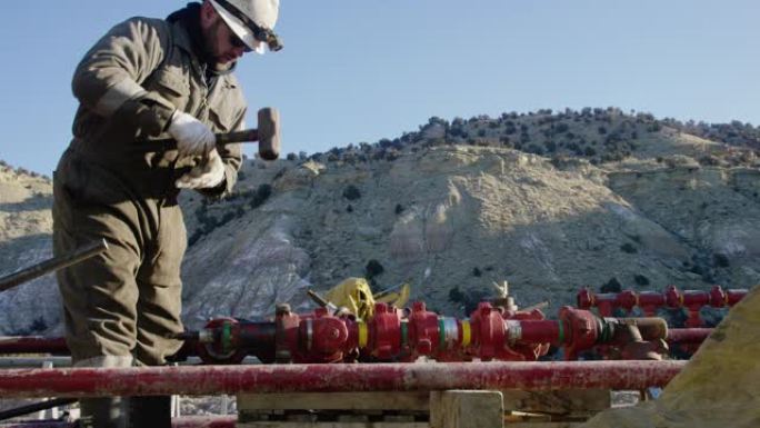 一个三十多岁的油田工人的慢动作镜头，在一个寒冷，阳光明媚的冬天的早晨，在石油和天然气钻井平台现场用锤