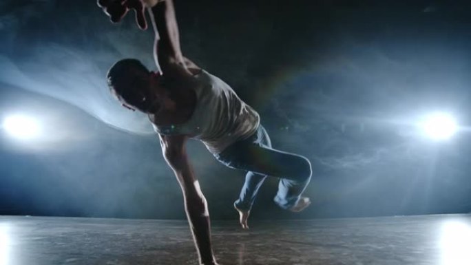 现代芭蕾舞剧，一个男人从舞台上的一个坐姿跳起翻筋斗，在聚光灯下的烟雾中，在黑暗的舞台上旋转到摄像机上