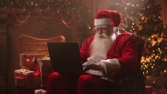 留着白胡子的圣诞老人坐在椅子上，在笔记本电脑上工作，阅读孩子们的电子邮件。