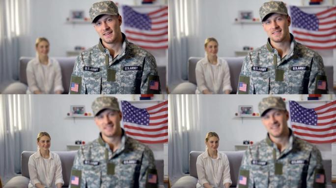 美国士兵对着镜头微笑，妻子自豪地坐在沙发上，背靠美国国旗
