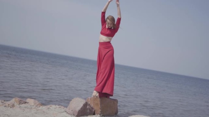 肖像优雅的年轻女子穿着红色长裙跳舞，举起双手在大石头上。
