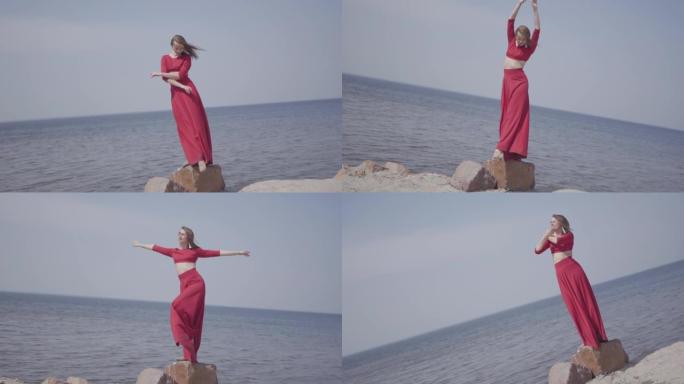 肖像优雅的年轻女子穿着红色长裙跳舞，举起双手在大石头上。