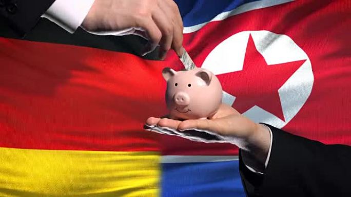 德国投资朝鲜，手把钱放在存钱罐里，旗帜背景