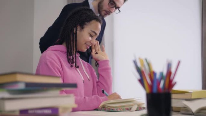 年轻的白人男老师来到非裔美国学生身边，翻阅她的笔记。漂亮害羞的女孩看着导师，用手指缠绕头发。爱，感情