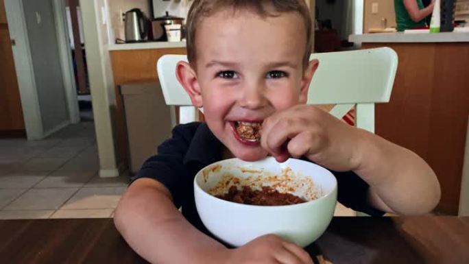 一个可爱的四岁白人男孩在吃饭时微笑，因为他的母亲在他身后的家庭厨房里工作