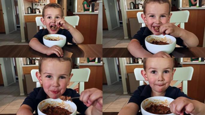 一个可爱的四岁白人男孩在吃饭时微笑，因为他的母亲在他身后的家庭厨房里工作