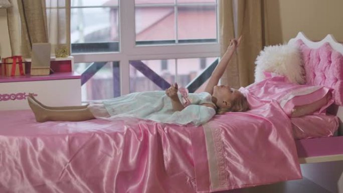 白人小女孩躺在房间的粉红色床上，打手势说话。积极的孩子周末在家休息。休闲活动，快乐童年。