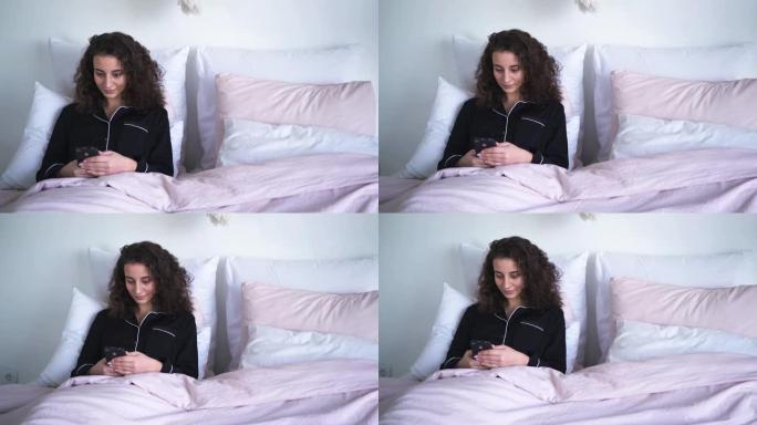 微笑的女人躺在床上，使用智能手机。可爱的黑发高加索女孩，卷发，早上检查社交媒体。