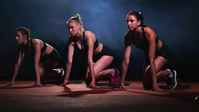 晚上在跑步机上穿着运动员黑色衣服的三个运动女孩将在距坐姿的冲刺距离处开始比赛